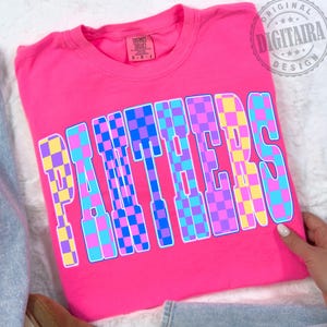 Puede incluir: Una camiseta rosa brillante con la palabra "PANTHERS" en letras grandes a cuadros. Las letras son una mezcla de cuadrados azules, morados, amarillos y rosas. La camiseta está doblada y parece ser de material suave.