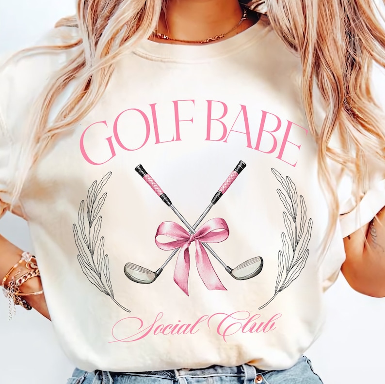 May include: A white t-shirt with the text "GOLF BABE Social Club" in pink lettering. The design features two crossed golf clubs with a pink bow.