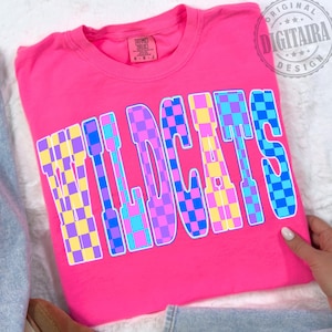 May include: A bright pink t-shirt with the word "WILDCATS" in large, checkered letters. The letters are filled with a pattern of blue, purple, and yellow squares. The shirt has a crew neck and short sleeves.