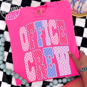 May include: A neon pink crewneck sweatshirt featuring the text OFFICE CREW in large, block-style letters. The letters are filled with a polka dot pattern in alternating shades of light pink, medium pink, and light blue with a white outline.