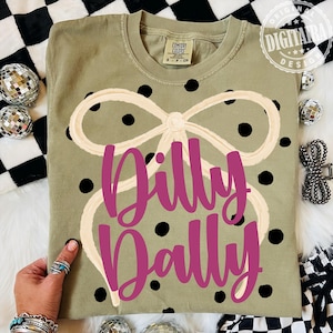 May include: Olive green t-shirt with a cream-colored bow design and black polka dots. The shirt features the magenta text "Dilly Dally". The shirt is a Comfort Colors brand.