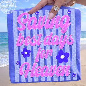 Peut inclure: Une serviette de plage à rayures bleues et blanches avec le texte "Saving best days for Heaven" en script rose. La serviette a des accents de fleurs bleues et est tenue par une main.