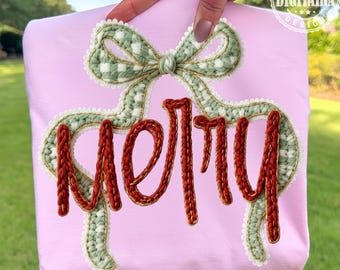 May include: A light pink sweatshirt with a green and white bow design and the word "Merry" in red, embroidered with a textured yarn. The design is accented with white lace trim. The sweatshirt is held up by a hand.