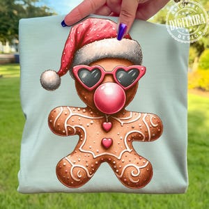 Op de afbeelding: Lichtblauw T-shirt met een peperkoekmannetje. Het peperkoekmannetje draagt een kerstmuts, hartvormige zonnebril en een roze kauwgomballenbel. Het ontwerp bevat decoratieve glazuurdetails en hartaccenten.