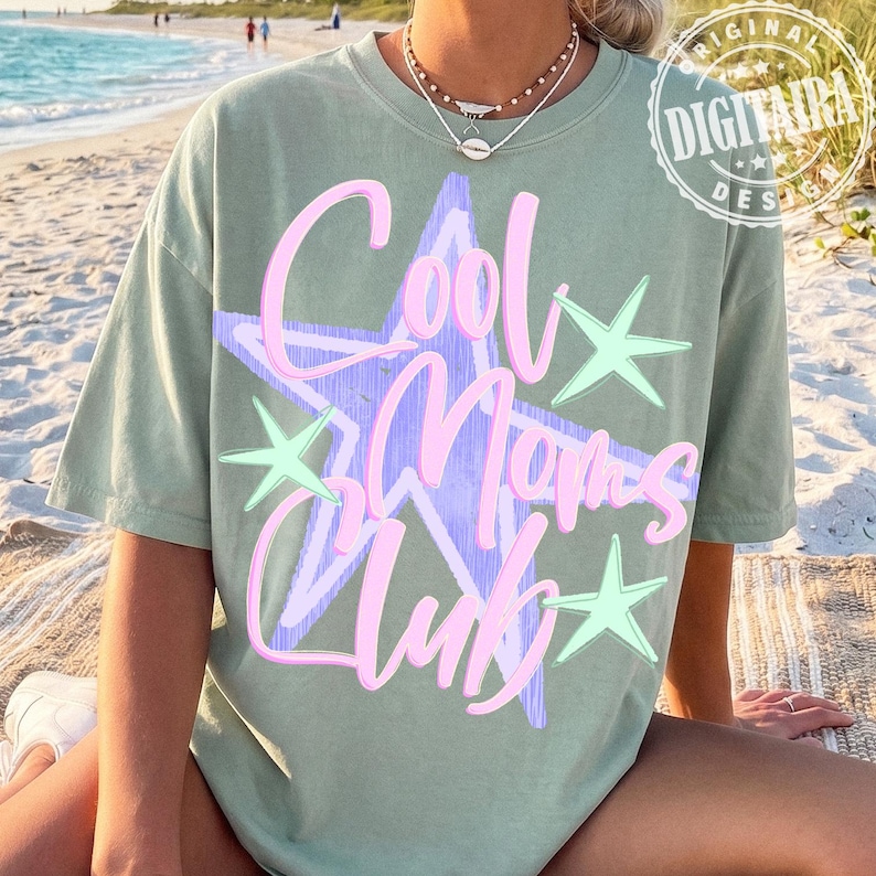 May include: Sage green t-shirt with the text "Cool Moms Club" in pink script over a light purple starburst design. The shirt also features light green star accents. The image is taken on a beach.
