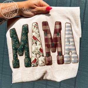 Könnte beinhalten: Ein cremefarbener Stoff mit dem Wort "MAMA" in verschiedenen Mustern appliziert. Die Buchstaben sind mit Schneeflocken, Kardinälen, Karos und Streifen verziert. Das Design wird von einer Hand mit einem rot lackierten Nagel gehalten.
