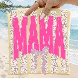 May include: A light yellow beach towel featuring the word "MAMA" in bright pink, distressed lettering. The towel has a vertical striped pattern with leopard print accents and a grey bow graphic. The towel is held up by a hand with gold rings.