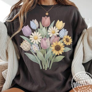 May include: Dark gray sweatshirt featuring a floral bouquet design. The bouquet includes tulips, daisies, and sunflowers in pastel colors and patterned fabrics. The design is stitched onto the shirt. The text "ORIGINAL DIGITAIRA DESIGN" is printed on the bottom right.