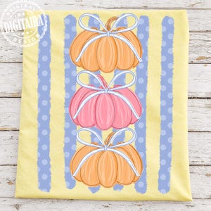 May include: A light yellow background with blue vertical stripes and polka dots. Three pumpkins are stacked vertically, each with a white bow. The top and bottom pumpkins are orange, and the middle pumpkin is pink.