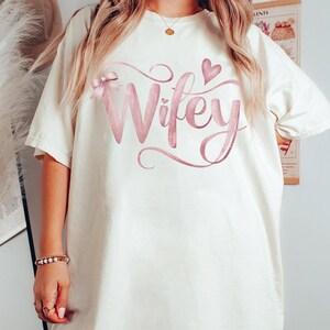 May include: White oversized t-shirt with a pink glitter design that says "Wifey" with a pink bow and heart.