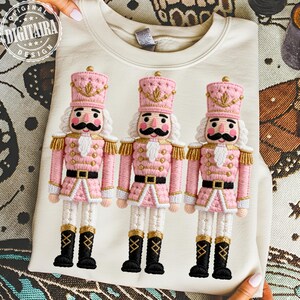 May include: Cream-colored sweatshirt featuring three embroidered pink and gold nutcracker figures. Each nutcracker has a black mustache, white hair, and black boots. The sweatshirt has the text "DIGITALRA DESIGN" in a circular logo.