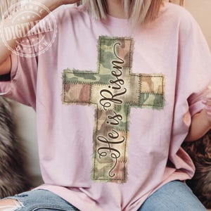 May include: A light pink t-shirt featuring a camouflage cross design with the words "He is Risen" in a cursive font. The cross is made of patches of green, brown, and tan fabric, stitched together with a visible seam.