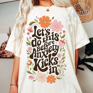 May include: White t-shirt with a floral design and the text "Let's do this before anxiety kicks in".