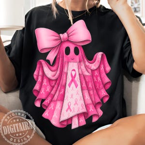 Puede incluir: Camiseta negra con un diseño de fantasma rosa con un gran lazo y detalles de cintas rosas. El fantasma está adornado con cintas rosas, que simbolizan la concienciación sobre el cáncer de mama. El diseño incluye el texto "ORIGINAL DIGITAIRA DESIGN".