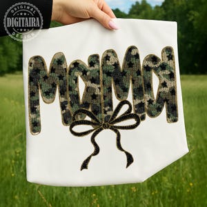 May include: A white tote bag with the word "MAMA" in a camouflage pattern with black stars. A black bow is embroidered below the word. The tote bag is held up by a person's hand in front of a green grassy background.