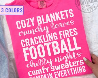 May include: A bright pink t-shirt with white text listing fall-themed words: "COZY BLANKETS, crunchy leaves, CRACKLING FIRES, FOOTBALL, chilly nights, comfy sweaters, PUMPKIN EVERYTHING." The shirt also has a "3 COLORS" label.