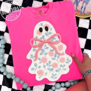 May include: A vibrant pink sweatshirt with a crocheted ghost motif. The ghost is white, adorned with pink and blue floral patterns, a pink bow, and black eyes. The sweatshirt has the text "DIGITALRA DESIGN" on the upper left.