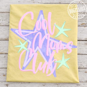 May include: Yellow t-shirt with the text "Cool Moms Club" in pink script over a purple starburst design. Mint green stars accent the design. The shirt has a relaxed fit.