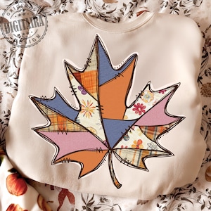 Op de afbeelding: Crèmekleurige sweatshirt met een groot, kleurrijk bladontwerp. Het blad bestaat uit verschillende stoffen patches in tinten oranje, blauw, roze en bloempatronen, omlijnd in bruin met gestikte details. Het ontwerp roept een herfstthema op.