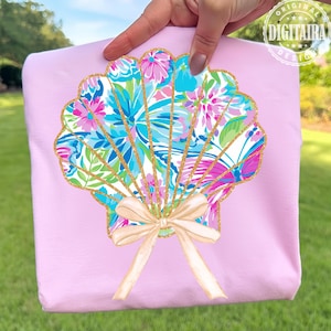 May include: A light pink tote bag featuring a seashell design. The seashell is filled with a colorful floral pattern in shades of blue, pink, and green, outlined with gold glitter. A beige bow is at the bottom of the seashell.