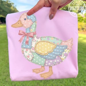 May include: A light pink fabric with a patchwork duck design. The duck features a pink head with a blue patch, a yellow beak, and a pink bow. The body is made of various floral and polka dot fabrics. The design is held by a hand.