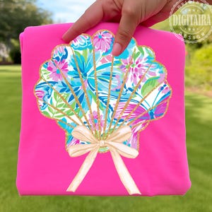 May include: A bright pink t-shirt featuring a seashell design. The seashell is filled with a floral pattern in shades of blue, pink, and green, outlined in gold glitter. A cream-colored bow is at the base of the shell.
