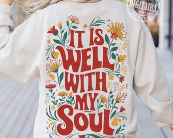 Puede incluir: Sudadera color crema con la frase "IT IS WELL WITH MY SOUL" en rojo, rodeada de ilustraciones florales en amarillo, rojo y verde. El diseño está centrado en la parte trasera de la prenda.