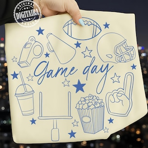 Puede incluir: Bolso tote color crema con ilustraciones azules de elementos temáticos de fútbol, incluyendo un balón, casco, palomitas de maíz y un dedo de espuma. Las palabras "Game day" están escritas en cursiva, rodeadas de estrellas.