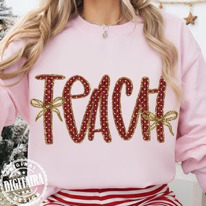 May include: A light pink sweatshirt with the word "TEACH" in a red, dotted design. The letters are outlined with a gold-colored thread and adorned with gold bows. The sweatshirt is part of a teacher-themed collection.