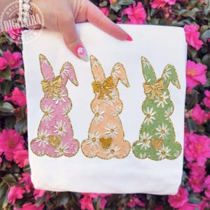 May include: White fabric featuring three Easter bunny designs. Each bunny is a different color: pink, peach, and green, decorated with white daisies, gold bows, and gold glitter trim. A gold heart is at the bottom of each bunny.