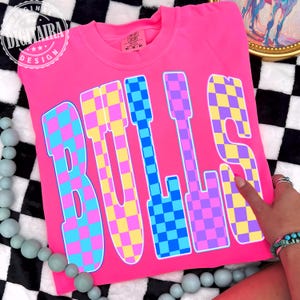 May include: A bright pink t-shirt with the word "BULLS" in large, checkered letters. The letters are a mix of blue, yellow, and purple squares. The shirt has a crew neck and short sleeves. The brand name "DIGITALRA DESIGN" is printed on the top left corner.