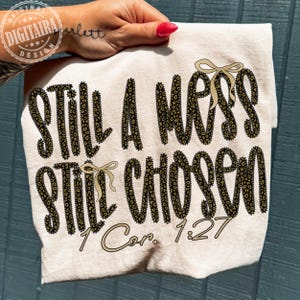 May include: Cream-colored t-shirt with the text "STILL A MESS STILL CHOSEN" in a leopard print design. The shirt also features a bow graphic and the Bible verse 1 Cor. 1:27.