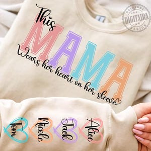 May include: A light beige sweatshirt with the text "This Mama wears her heart on her sleeve" in rainbow colours. The sleeve has four hearts with the names "Tom", "Nicole", "Jack", and "Alice" written inside.