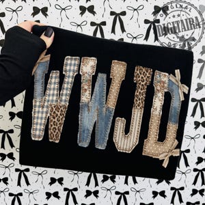 May include: Black clutch with the letters "WWJD" appliqued on the front. The letters are made of various patterned fabrics, including denim, leopard print, and floral designs. The clutch is held by a person with black nail polish.
