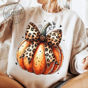 May include: A white t-shirt with a graphic of an orange pumpkin with a brown and black leopard print bow. The pumpkin has a stem and leaves.