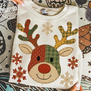 May include: A cream-coloured sweatshirt with a reindeer design. The reindeer has patchwork antlers and face, a red nose, black eyes, and a smiling mouth. Snowflakes surround the reindeer. The sweatshirt has the text "DIGITAIRA DESIGN" in a circular logo.
