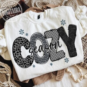 May include: White sweatshirt with the words "COZY season" in a mix of patterns, including leopard print, stars, and a checkered design. The design is accented with small blue snowflakes.