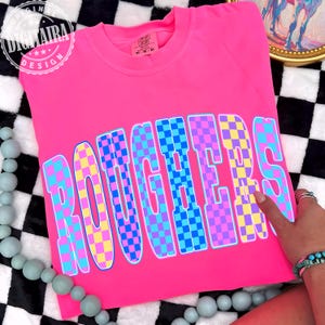 May include: A bright neon pink crewneck sweatshirt featuring the word ROUGHERS in large, bold, block letters. Each letter is filled with a vibrant checkerboard pattern in alternating shades of yellow, purple, blue, and cyan with a white outline.