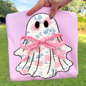 May include: A pale pink sweatshirt with a crochet ghost motif. The ghost is white, adorned with pink roses and blue leaves, black eyes, and a pink bow. The design is centrally placed on the sweatshirt.