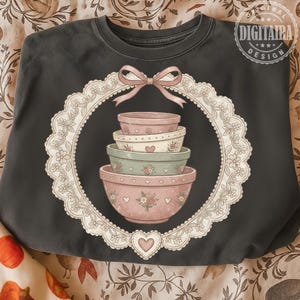 Puede incluir: Sudadera gris oscuro con un diseño decorativo. El diseño incluye una pila de cuencos para mezclar de colores pastel dentro de un círculo con ribete de encaje blanco, rematado con un lazo rosa. Los cuencos están decorados con corazones y motivos florales. El texto "DIGITAIRA DESIGN" está en un círculo en la parte superior derecha.