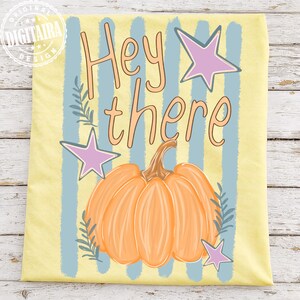 May include: A yellow t-shirt with a pumpkin illustration and the words "Hey there". The design includes light blue vertical stripes, pink stars, and green leaf accents, creating a fall theme.