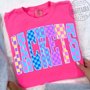 May include: A bright pink t-shirt with the word "JACKETS" in large, colorful checkered letters. The letters feature a pattern of blue, purple, yellow, and pink squares. The shirt is folded and appears to be made of soft material.