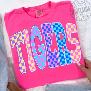 May include: A bright pink t-shirt with the word "TIGERS" in large, checkered letters. The letters are a mix of blue, pink, yellow, and purple squares. The shirt has a crew neck and short sleeves. The shirt is made of a soft material.