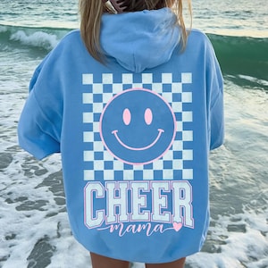 Op de afbeelding: Lichtblauwe hoodie met een smiley-gezicht op de achterkant. Het smiley-gezicht staat in een roze omkaderde vierkant met een dambordpatroon. De tekst "CHEER mama" staat eronder in wit en lichtblauw.