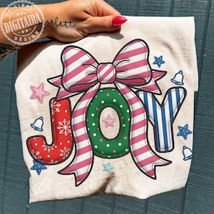 以下が含まれることがあります： ベージュ色のTシャツ。お祝いの「JOY」のデザイン。ピンクと白のストライプのリボン、白い水玉模様の緑色のリース、赤、緑、青の文字が含まれています。装飾的な星とベルがテキストを囲んでいます。