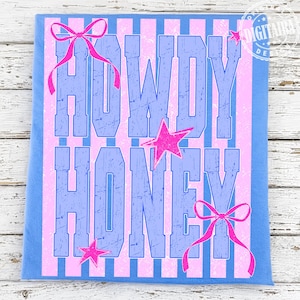 May include: A blue t-shirt with a pink and blue striped design. The words "HOWDY HONEY" are printed in distressed blue lettering, with pink bows and stars. The shirt is on a white wooden surface.