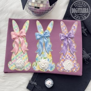 May include: A purple decorative piece featuring three bunny figures with pastel floral and plaid patterns. Each bunny has a large, textured bow in pink, blue, and purple. White floral accents are at the base of each bunny.