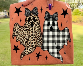May include: An orange-colored Halloween-themed graphic featuring two ghost figures. One ghost has a leopard print design, and the other has a black and white checkered pattern. Both ghosts wear black lace bows. Black stars and diamond shapes surround the ghosts.