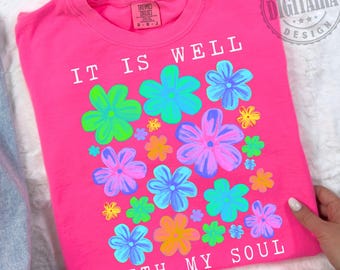 May include: A bright pink t-shirt with the words "IT IS WELL WITH MY SOUL" in white. The shirt features a floral design with flowers in shades of green, blue, purple, and yellow. The shirt is a crew neck style.