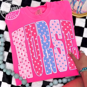 May include: A bright pink t-shirt with the word "NURSE" in large, polka-dotted letters. The letters are outlined in white, with pink and blue polka dots. The shirt is a Comfort Colors brand.